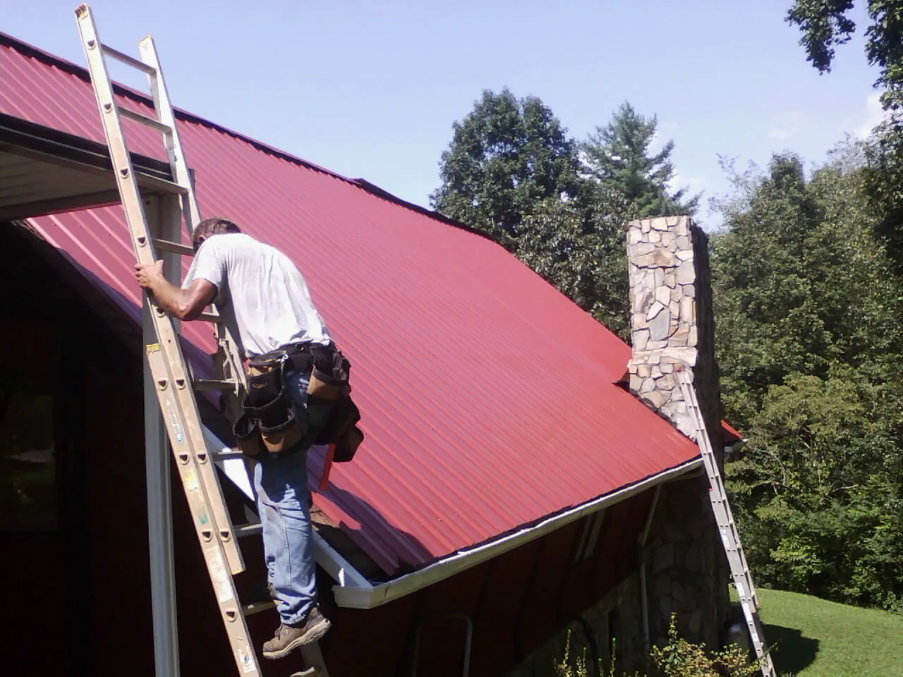 Roofer on a ladder beside a red metal roof and stone chimney — Soffit & Fascia Repair by Roofing Brothers in Proctorsville